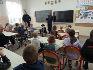 Policjanci w klasie  wszkole stojący przed tablicą przed grupą dzieci siedzących w szkolnych ławkach
