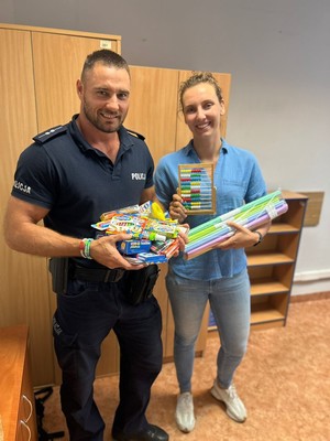 Umundurowany policjant oraz nie umundurowana policjantka stojący razem w dłoniach trzymający przybory szkolne