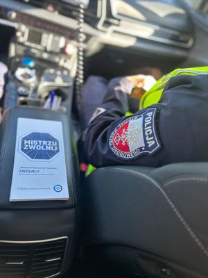Policjant siedzący w radiowoze, obok niego na podłokietniku ulotka Mistrzu, zwolnij!