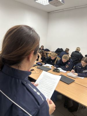 Policjantka w szkolnej sali stojąca z kartką w dłoni przed grupą uczni klasy mundurowej