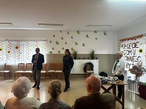 Policjantka z pracownikami Zus stojąca w sali przed seniorami siedzącymi wokól na krzesłach