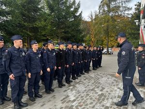 Policjant stojący przed budynkiem szkoły, przed nim w szeregu stoją uczniowie klasy mundurowej