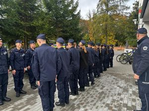 Policjant stojący przed budynkiem szkoły, przed nim w szeregu stoją uczniowie klasy mundurowej