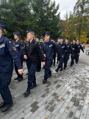 Policjant stojący przed budynkiem szkoły, przed nim w szeregu stoją uczniowie klasy mundurowej