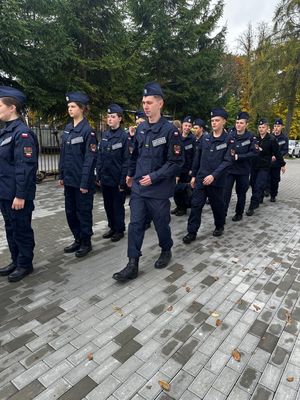 Policjant stojący przed budynkiem szkoły, przed nim w szeregu stoją uczniowie klasy mundurowej