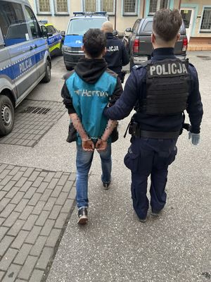 Policjant prowadzący zatrzymanego przez plac wewnętrzy komendy, wokół stoją policyjne radiowozy.