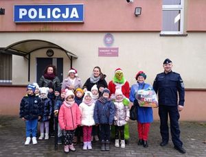 Policjant z dziećmi stojący przed budynkiem komisariatu Policji pozujacy do zdjęcia.