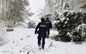 Policjanci idący przez las w zimowej sceneri przed nim stoi radiowóz